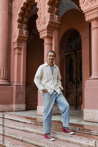 full length of pleased african american man in shirt jacket standing with hands in pockets near building in europe.