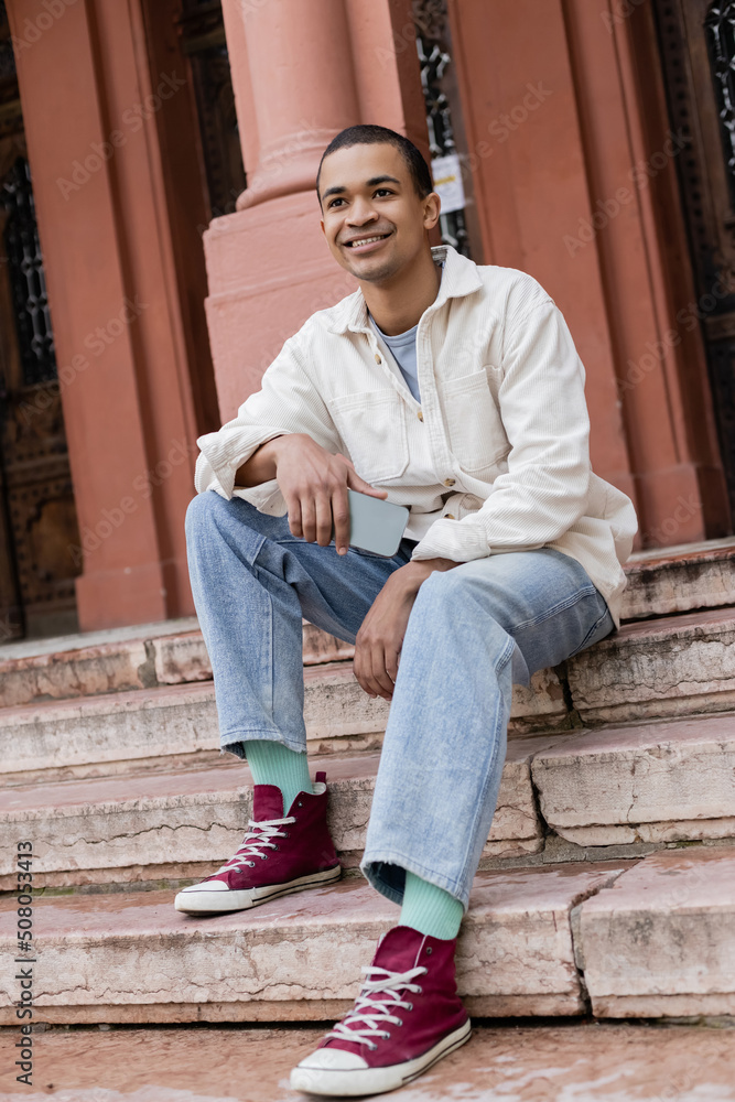 full length of smiling african american man in shirt jacket holding smartphone while sitting on stairs in city. full length of smiling african american man in shirt jacket holding smartphone while sitting on stairs in city.