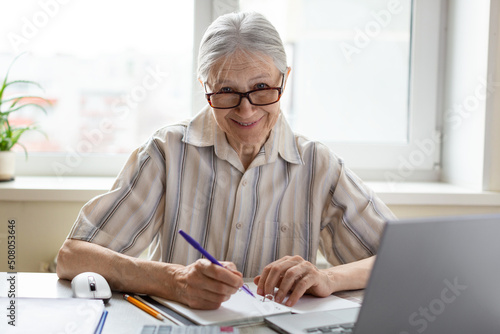 Friendly smiling caucasian senior woman in eyeglasses sits at the laptop with a pen in hand at home, selective focus.