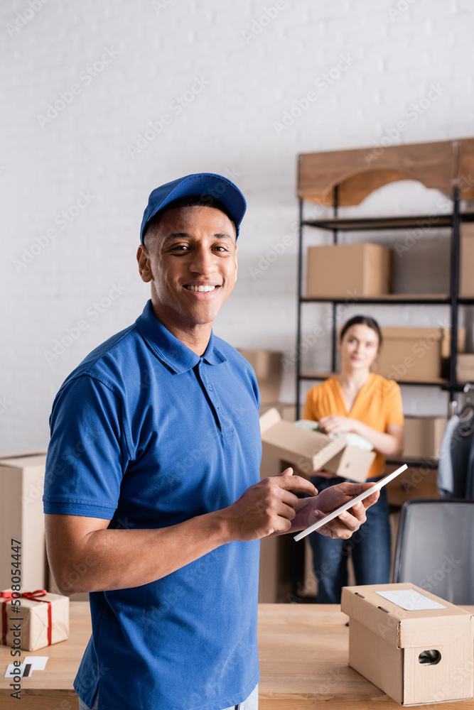 Smiling african american deliveryman holding digital tablet near boxes in online web store.