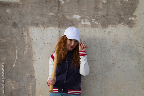young and beautiful red-haired woman with baseball cap, jacket and glove with baseball bat resting on her shoulder on grey cement background. Sport and recreation concept. © @skuder_photographer