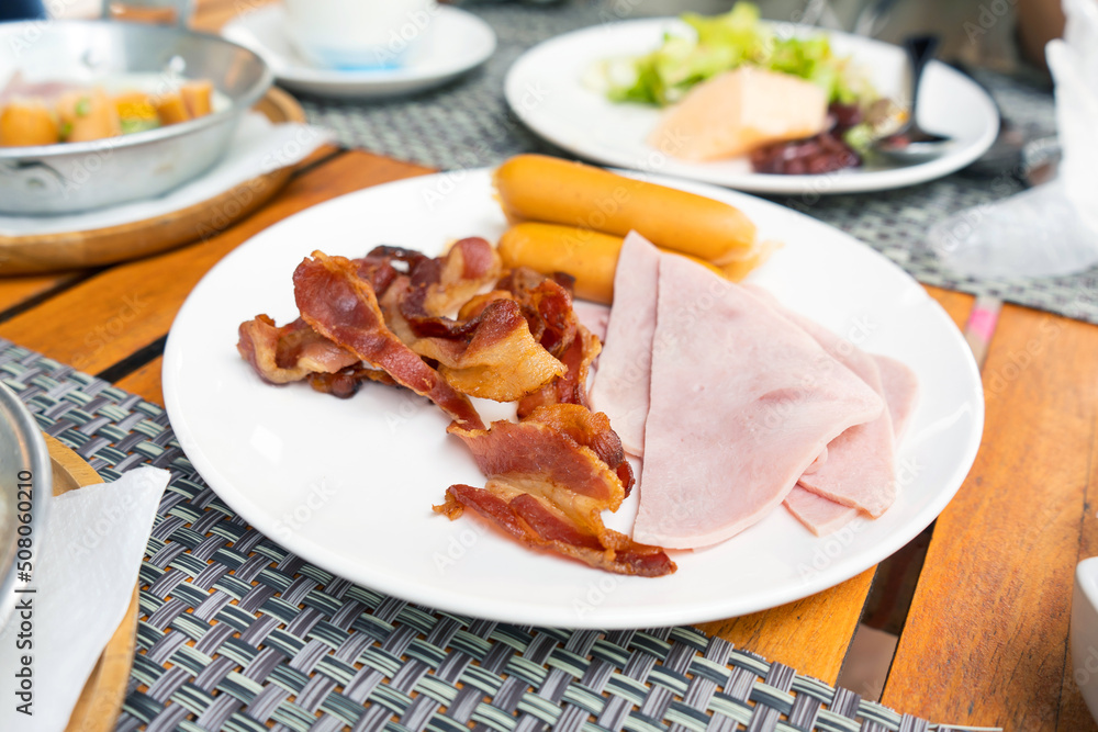 The breakfast menu consisted of ham, sausage, bacon in a white plate on the table.