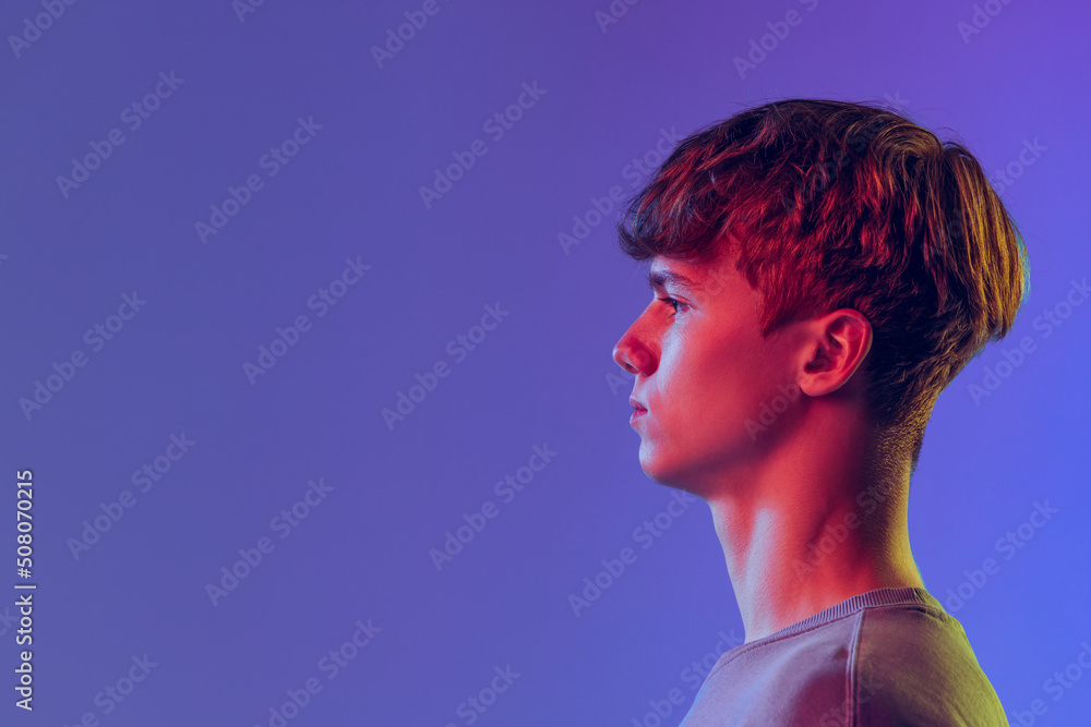 Profile view young red-headed man, student isolated over blue studio background in neon light. Concept of youth, fashion, emotions, facial expression
