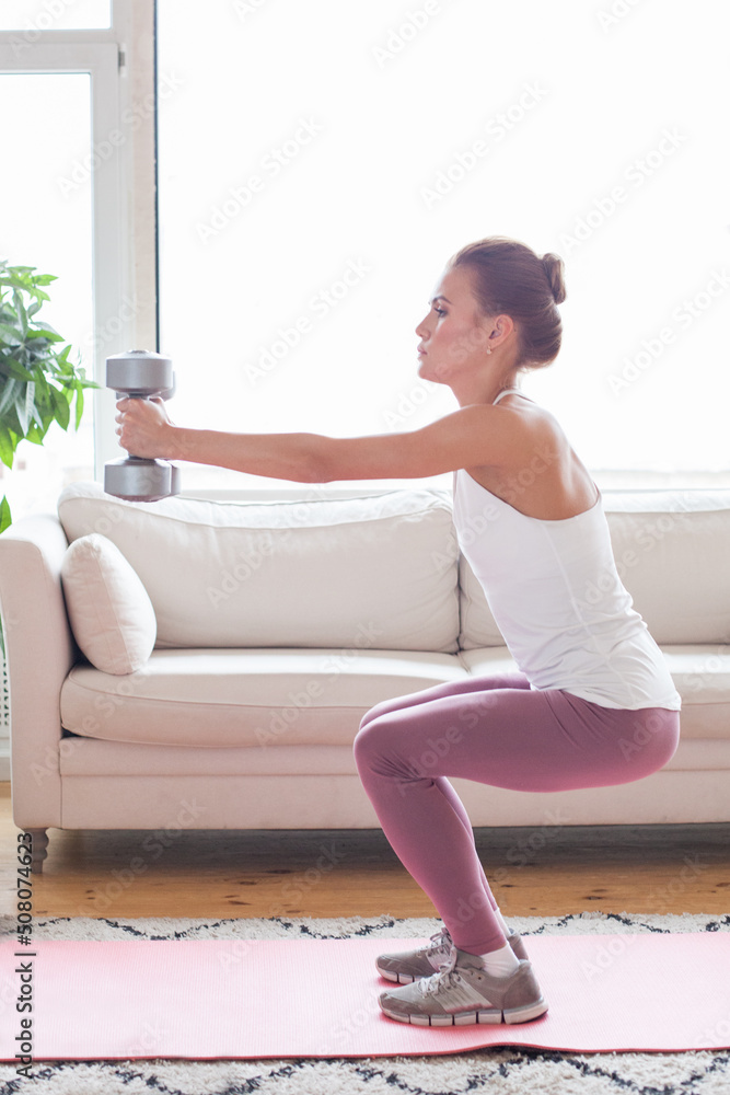 Young adult woman doing fitness exercises with gray dumbbells at her home in living room Young adult woman doing fitness exercises with gray dumbbells at her home in living room