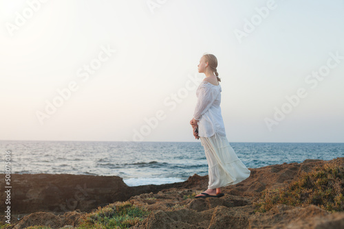Pensive teenager girl in white dress standing on the beach looking away at the horizon in the morning