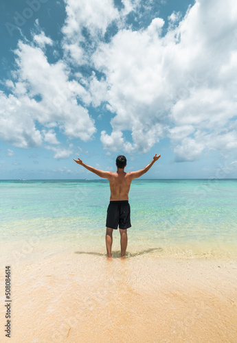 young hispanic man from the back raises his arms in front of the caribbean sea
