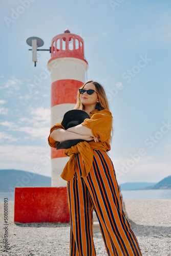 Fashionable woman on vacation against the backdrop of the sea, mountains and a lighthouse. Summer, sunny day, walk along the beach, enjoy life and travel. photo