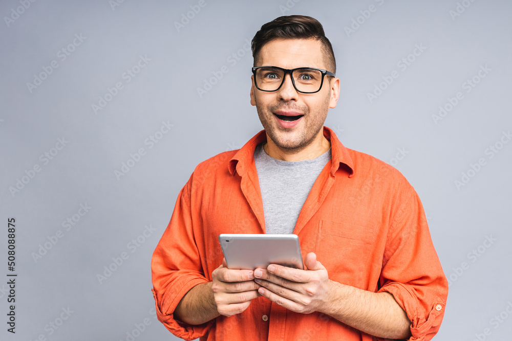 Shocked amazed young ukrainian man in casual standing and using tablet isolated over grey white background. Shocked amazed young ukrainian man in casual standing and using tablet isolated over grey white background.