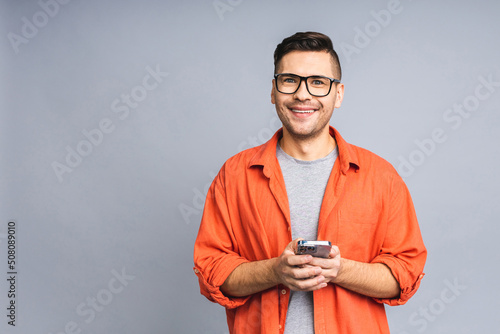 Great news today! Happy smiling young ukrainian man in casual standing and using mobile phone isolated over grey white background. © denis_vermenko
