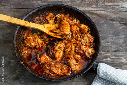Pieces of baked chicken in sauce in a frying pan with a wooden spatula on a dark table.