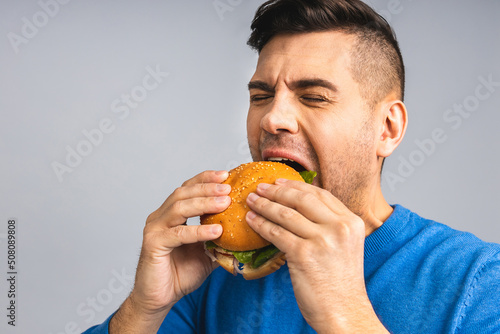 Young ukrainian man holding a piece of hamburger. Student eats fast food. Burger is not helpful food. Very hungry guy. Diet concept. Isolated over grey white background.