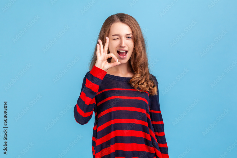 Portrait of woman wearing striped casual style sweater standing showing ok sign and winking to camera, approval sign, agreeing with suggestion. Indoor studio shot isolated on blue background. Portrait of woman wearing striped casual style sweater standing showing ok sign and winking to camera, approval sign, agreeing with suggestion. Indoor studio shot isolated on blue background.