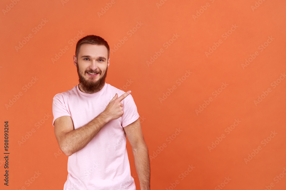 Portrait of bearded man standing, looking at camera with happy positive face and pointing at wall copyspace, wearing pink T-shirt. Indoor studio shot isolated on orange background. Portrait of bearded man standing, looking at camera with happy positive face and pointing at wall copyspace, wearing pink T-shirt. Indoor studio shot isolated on orange background.