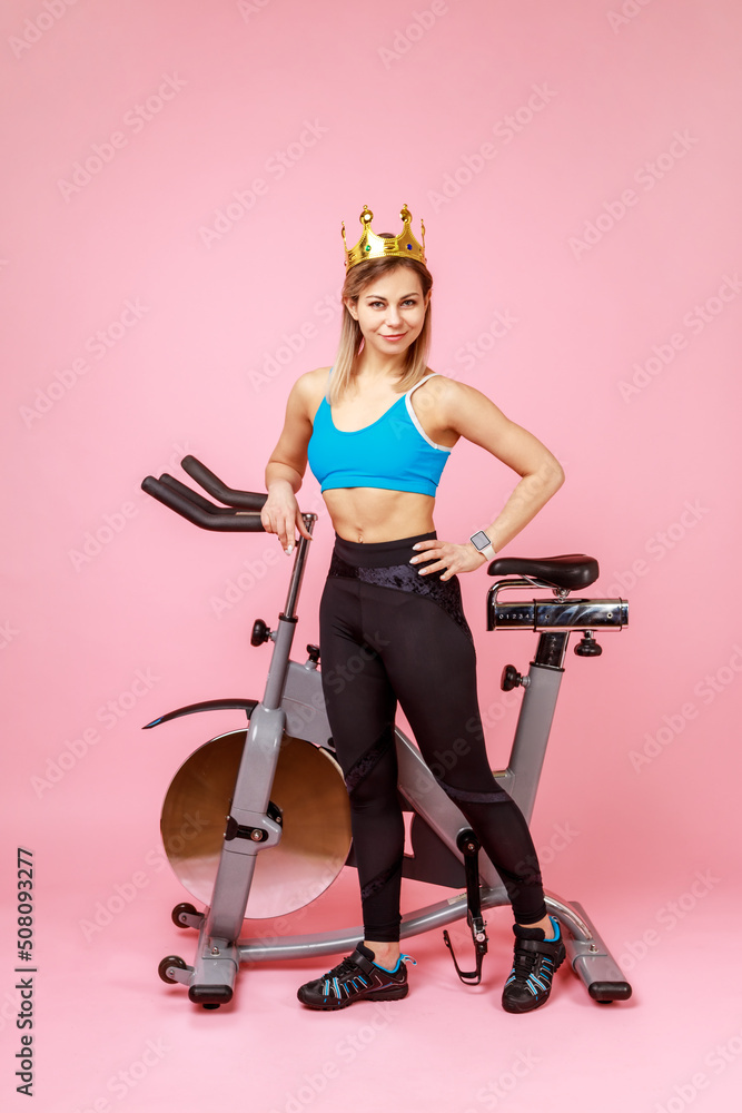 Full length of athletic woman in golden crown, queen of sports trainings, standing with hand on hips near bike simulator, wearing sports tights and top. Indoor studio shot isolated on pink background.