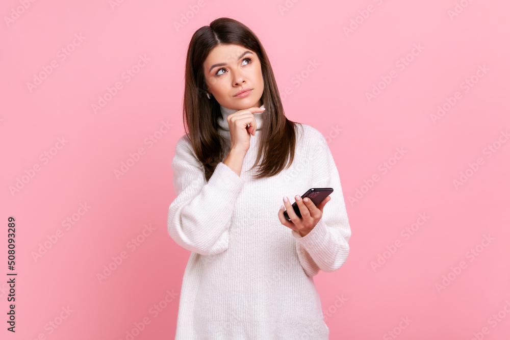 Thoughtful beautiful young adult brunette girl holding smart phone in hands, looking away, thinking, wearing white casual style sweater. Indoor studio shot isolated on pink background. Thoughtful beautiful young adult brunette girl holding smart phone in hands, looking away, thinking, wearing white casual style sweater. Indoor studio shot isolated on pink background.