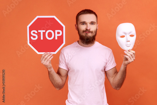 Portrait of puzzled confused bearded man holding white mask with unknown face and red traffic sign, looking at camera, wearing pink T-shirt. Indoor studio shot isolated on orange background. photo