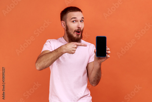 Portrait of bearded man pointing at cellphone and with amazed expression, recommending gadget or mobile application, wearing pink T-shirt. Indoor studio shot isolated on orange background. © khosrork