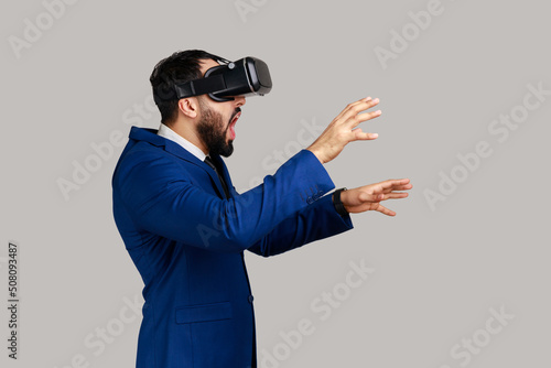 Side view of man in vr headset, playing virtual reality game with shocked facial expression, outstretching hands, wearing official style suit. Indoor studio shot isolated on gray background.