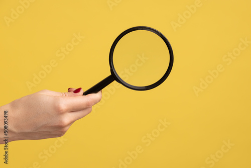 Profile side view closeup of woman hand holding magnify glass, loupe. Indoor studio shot isolated on yellow background.