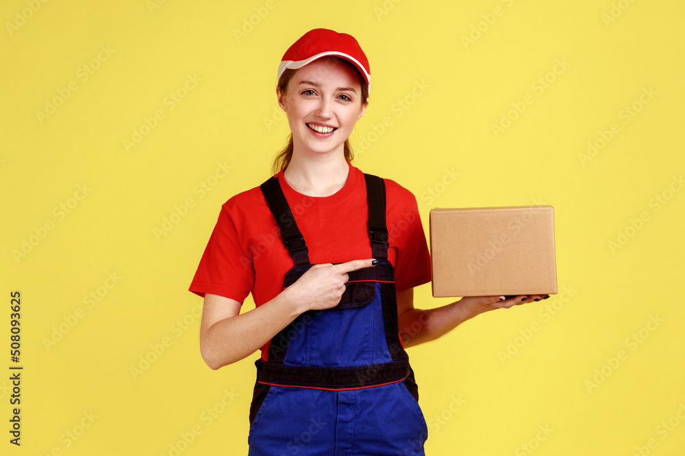 Positive smiling courier woman standing and pointing at cardboard parcel, online order for client, looking at camera, wearing overalls and red cap. Indoor studio shot isolated on yellow background.