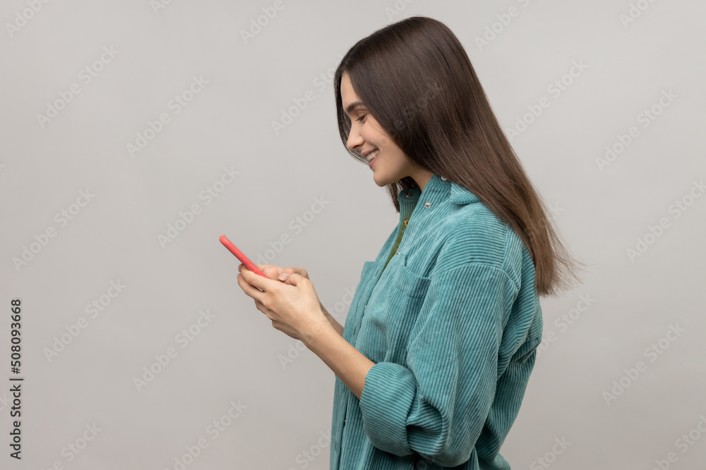 Side view of happy woman reading message on smartphone and smiling, using mobile device for communication, browsing web, wearing casual style jacket. Indoor studio shot isolated on gray background. Side view of happy woman reading message on smartphone and smiling, using mobile device for communication, browsing web, wearing casual style jacket. Indoor studio shot isolated on gray background.