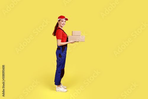Side view portrait of courier woman standing with two parcels in hands, looking at camera, delivery service, wearing overalls and red cap. Indoor studio shot isolated on yellow background.