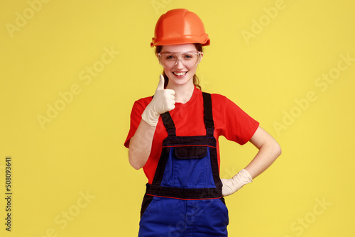 Portrait of positive worker woman standing looking at camera with optimistic expression, showing thumb up, wearing overalls and helmet. Indoor studio shot isolated on yellow background. © khosrork