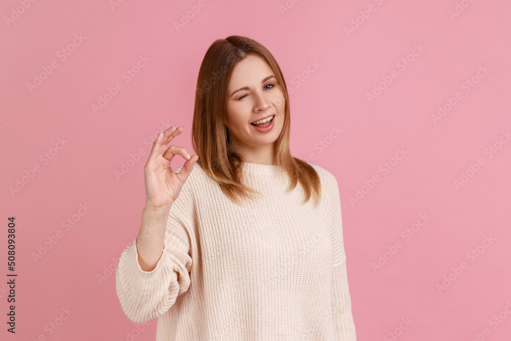 Portrait of attractive optimistic blond woman looking at camera showing ok sign gesture and winking, wearing white sweater. Indoor studio shot isolated on pink background. Portrait of attractive optimistic blond woman looking at camera showing ok sign gesture and winking, wearing white sweater. Indoor studio shot isolated on pink background.