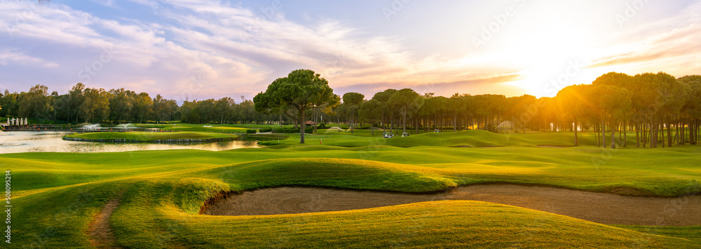 Golf course at sunset with beautiful sky and sand trap. Scenic panoramic view of golf fairway with bunker. Golf field with pines - obrazy, fototapety, plakaty 