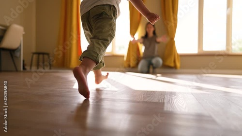 Happy family. Baby feet walk on floor towards mother. Child makes steps barefoot on parquet floor in children room. Son runs to his mother on the laminate with his bare feet. happy baby first steps