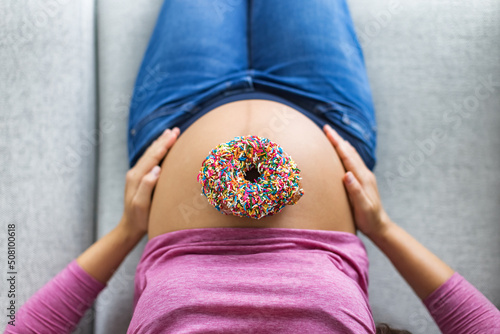 Pregnant woman with donut on belly top view. Cravings of desserts and sweets during pregnancy, Pastry with birthday cake sprinkles on baby bump for gestational diabetes photo