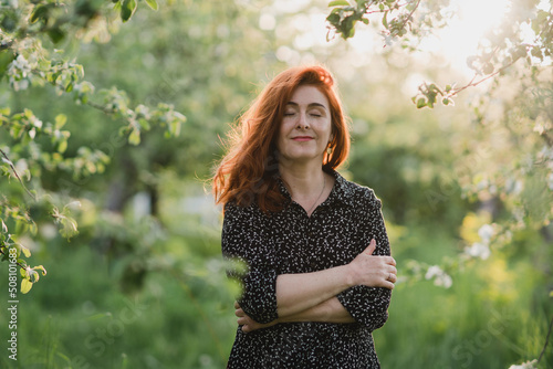 Portrait of gorgeous middle aged woman in casual dress relaxing in a blooming spring garden. Happy senior woman smiling and looking at camera. Red-haired mature lady posing outdoors in a spring day.