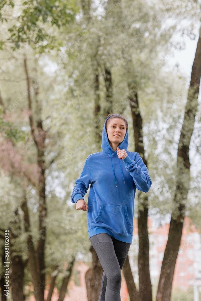 a European woman in a blue hoodie is running and doing sports in the park. selective focus, fitness and yoga outdoor sports, exercise and meditation and fitness for body health. spiritual development a European woman in a blue hoodie is running and doing sports in the park. selective focus, fitness and yoga outdoor sports, exercise and meditation and fitness for body health. spiritual development