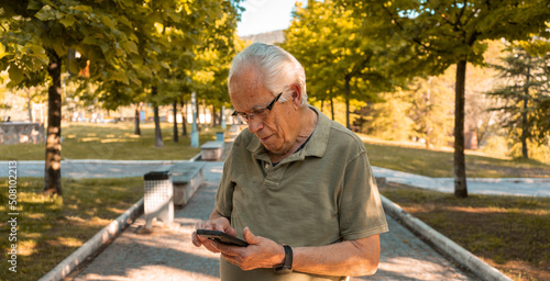 Hombre mayor en el parque, mirando su teléfono móvil con expresión de preocupación. Fotografía horizontal con espacio para texto. photo
