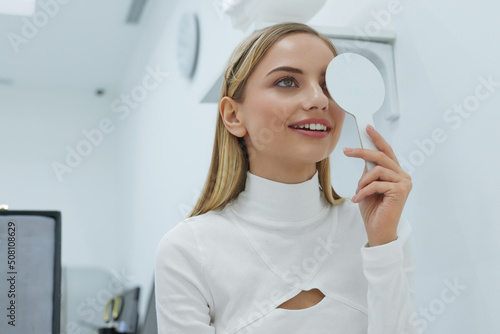 Eye Exam. Smiling Young Woman Doing Eye Test in Ophthalmological Clinic  Holding Occluder and Looking at Chart. Eyesight correction in Optics Clinic Concept