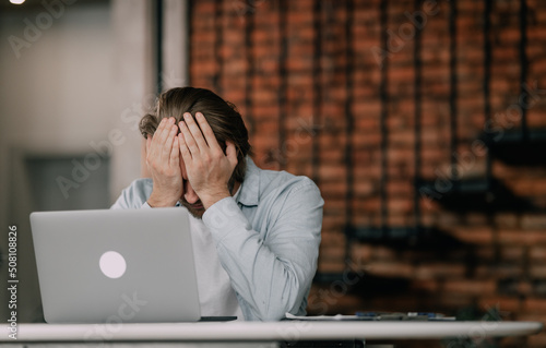 Stressful office job finally turns young man into a burnout. Stressed, tired, exhausted male worker overwhelmed with problems sitting at working desk with laptop computer and holding hand on his face