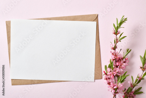 The close-up layout consists of a kraft envelope with blank paper and a delicate sprig of pink flowers on a pastel pink background. © Irina Lesovaia