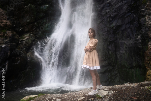 Lonely girl in a short beige dress stands next to the waterfall.