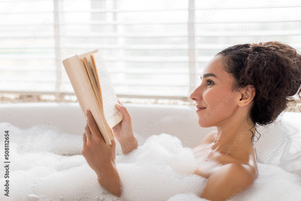 Beautiful young woman lying in foamy bath, reading book, enjoying fascinating story in bathtub at home, copy space Beautiful young woman lying in foamy bath, reading book, enjoying fascinating story in bathtub at home, copy space