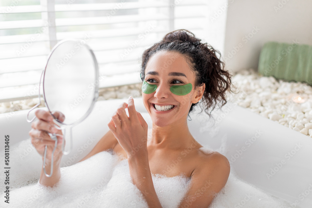 Sexy young woman lying in bubble bath, applying eye patches near mirror at home, free space. Domestic spa concept Sexy young woman lying in bubble bath, applying eye patches near mirror at home, free space. Domestic spa concept