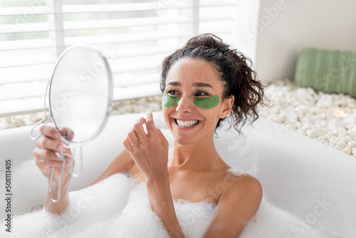 Sexy young woman lying in bubble bath, applying eye patches near mirror at home, free space. Domestic spa concept