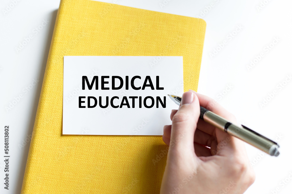 Text MEDICAL EDUCATION text on a card that lies on a yellow notepad on a table next to a person's hand with a pen Text MEDICAL EDUCATION text on a card that lies on a yellow notepad on a table next to a person's hand with a pen