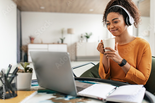 Smiling black lady watching webinar on computer, drinking hot coffee