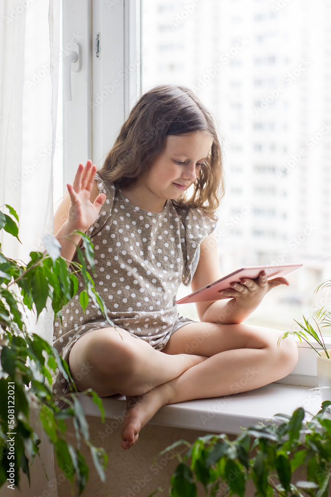 Positive caucasian little girl sitting near window at home with her digital tablet, chatting or have video call. Positive caucasian little girl sitting near window at home with her digital tablet, chatting or have video call.