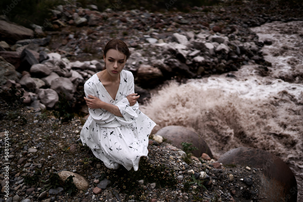Sensual elegant girl with brown hair in a white dress sits on the stone river's beach in the mountains. Incredible nature and attractive girl as wallpaper or background. Sensual elegant girl with brown hair in a white dress sits on the stone river's beach in the mountains. Incredible nature and attractive girl as wallpaper or background.
