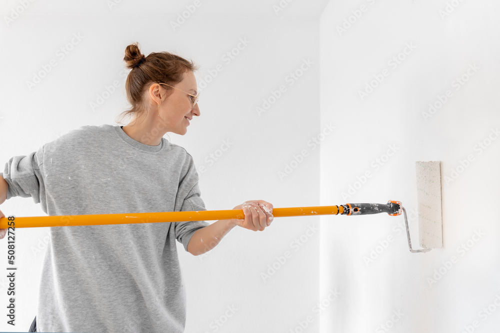 Portrait young adult beautiful single happy woman painting wall paint roller brush white color living room moving new home flat . Female painter decorating interior renovation residential apartment Portrait young adult beautiful single happy woman painting wall paint roller brush white color living room moving new home flat . Female painter decorating interior renovation residential apartment