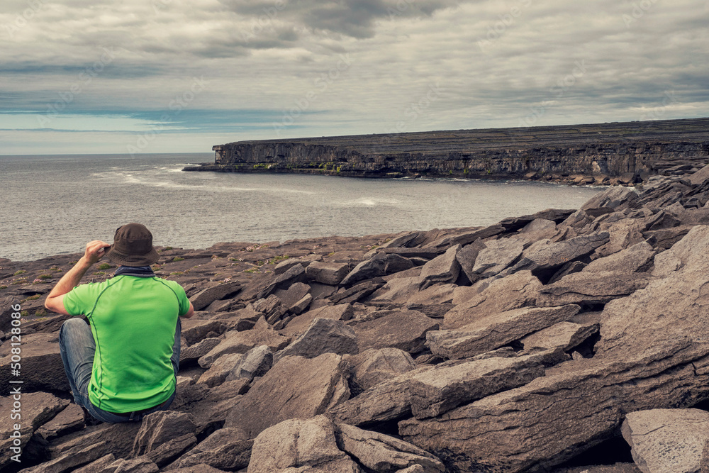 Male tourist on a rough stone surface of a cliff. Ocean and dramatic cloudy sky in the background. Aran island, Ireland. Stunning Irish landscape. Travel and tourism concept.