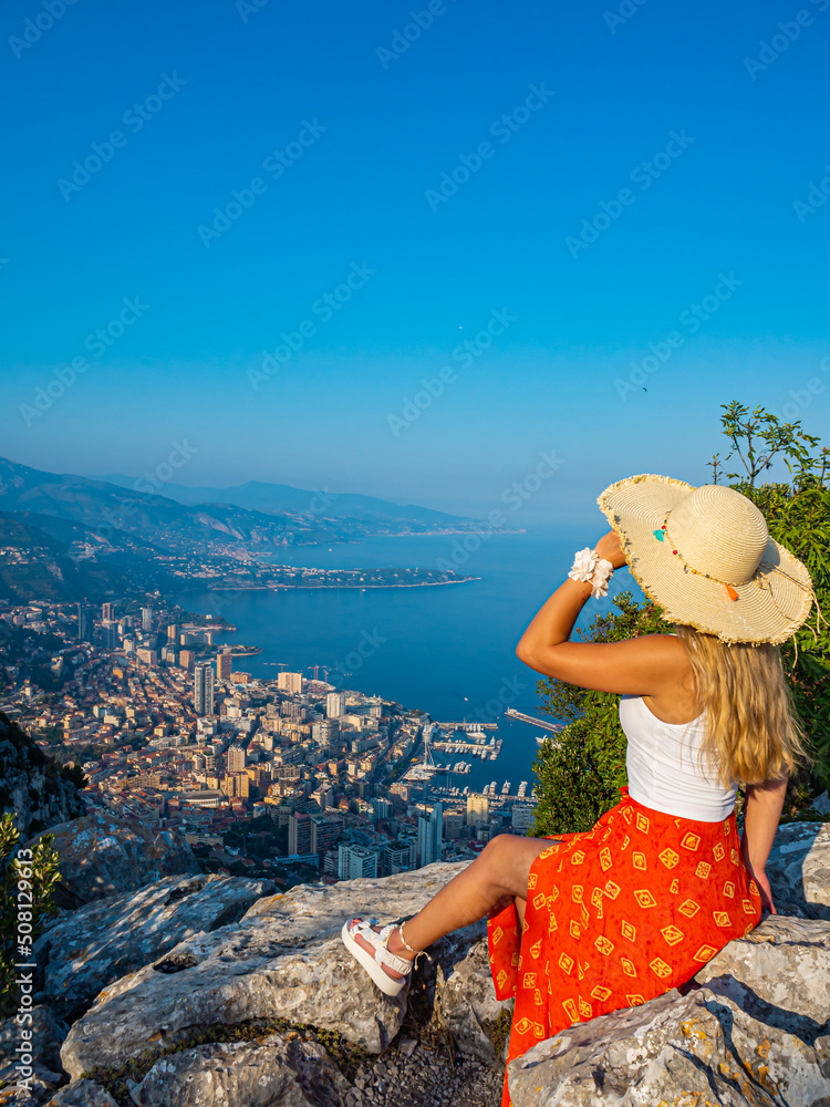 Young blonde hair girl in a hat sitting on the edge of a mountain with a beautiful panoramic view of Monaco during the setting sun, Monte Carlo, Cote d'azur