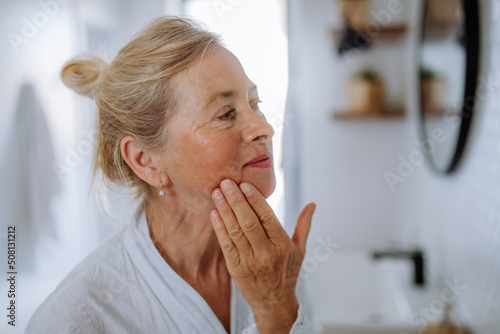 Beautiful senior woman in bathrobe looking at mirror and applying natural face cream in bathroom, skin care concept.