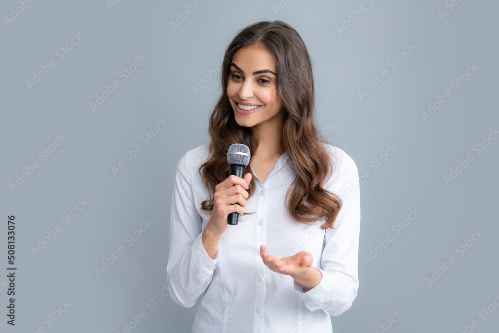 Beautiful business woman is speaking on conference. Happy smiling business woman holding mic, standing with microphone against grey background, wearing shirt. Beautiful business woman is speaking on conference. Happy smiling business woman holding mic, standing with microphone against grey background, wearing shirt.
